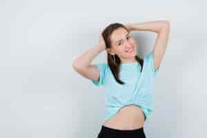 Foto gratuita chica joven con las manos detrás de la cabeza en camiseta turquesa, pantalones y mirando feliz, vista frontal.