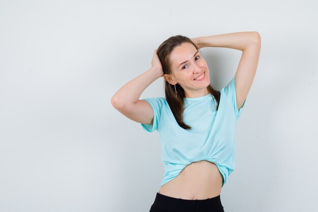 Chica joven con las manos detrás de la cabeza en camiseta turquesa, pantalones y mirando feliz, vista frontal.