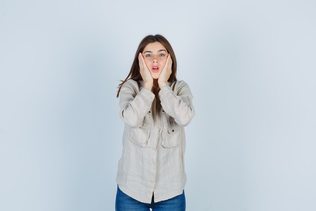 Chica joven con las manos en las mejillas, manteniendo la boca abierta en camisa beige, jeans y mirando sorprendido, vista frontal.