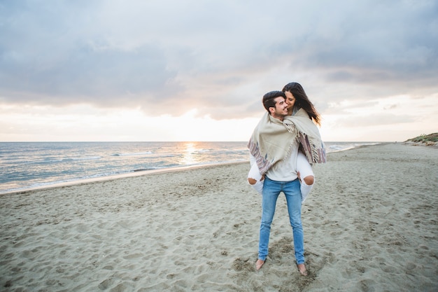 Chica joven con una manta subida a la espalda de su novio en la playa