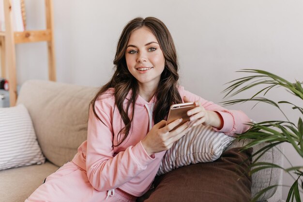 Chica joven con un maquillaje suave y un hermoso corte de pelo y con un teléfono inteligente en las manos sonríe alegremente Una mujer genial vestida de rosa se divierte en casa los fines de semana