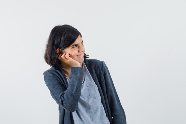 Chica joven con la mejilla apoyada en la palma de la mano, mirando a la derecha con una camiseta gris claro y una sudadera con capucha frontal con cremallera gris oscuro y un aspecto lindo