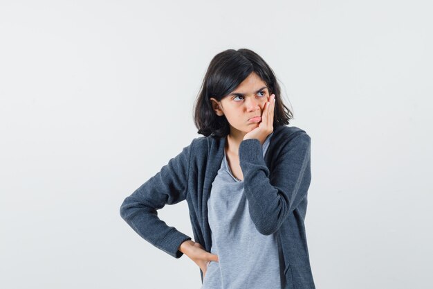 Chica joven con la mejilla apoyada en la palma de la mano, mirando a otro lado con una camiseta gris claro y una sudadera con capucha gris oscuro con cremallera y mirando pensativa.