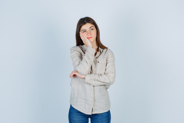 Chica joven con la mejilla apoyada en la palma, pensando en algo en camisa beige, jeans y mirando pensativo, vista frontal.