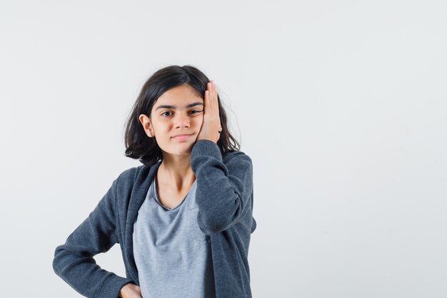 Chica joven con la mejilla apoyada en la palma, poniendo la mano en la cintura con una camiseta gris claro y una sudadera con capucha frontal con cremallera gris oscuro y un aspecto lindo,