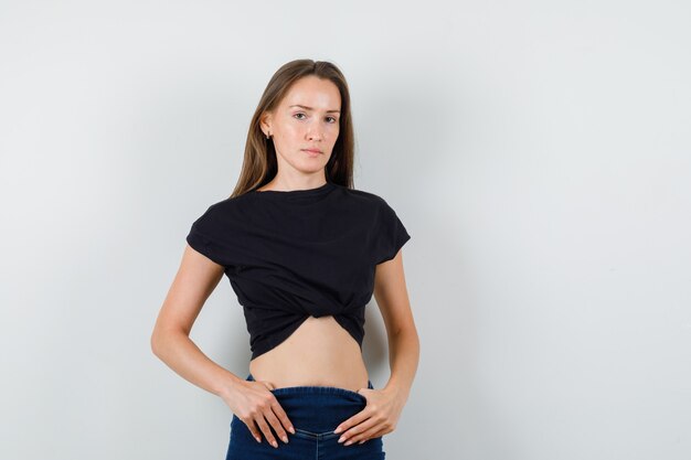 Chica joven mirando a la cámara en blusa negra, pantalones y mirada seria.