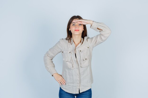 Chica joven mirando lejos con las manos sobre la cabeza, con la mano en la cadera en camisa beige, jeans y luciendo linda. vista frontal.