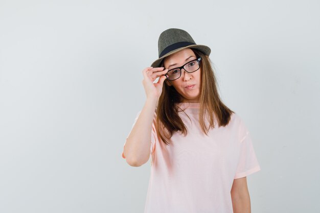 Chica joven mirando a través de gafas en camiseta rosa, sombrero y mirando indeciso, vista frontal.