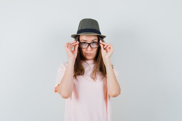 Chica joven mirando a través de gafas en camiseta rosa, sombrero y mirando vacilante. vista frontal.