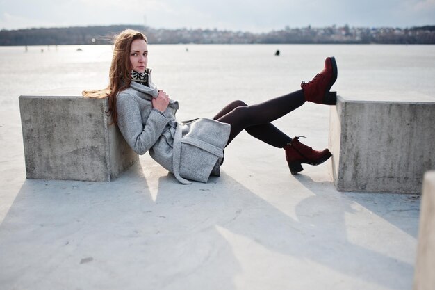 Chica joven modelo en abrigo gris posó sentada cerca del cubo de piedra contra el lago congelado