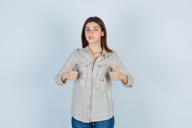 Chica joven mostrando doble pulgar hacia arriba en camisa beige, jeans y mirando seria, vista frontal.