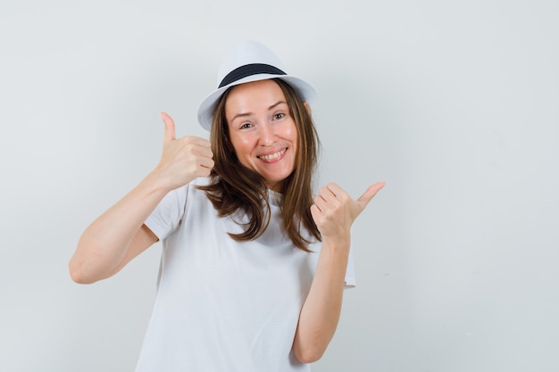 Chica joven mostrando doble pulgar hacia arriba en camiseta blanca, sombrero y mirando feliz, vista frontal.