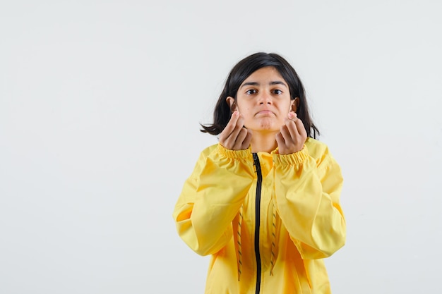 Chica joven mostrando gesto de dinero y apretando el puño en chaqueta de bombardero amarilla y mirando serio.