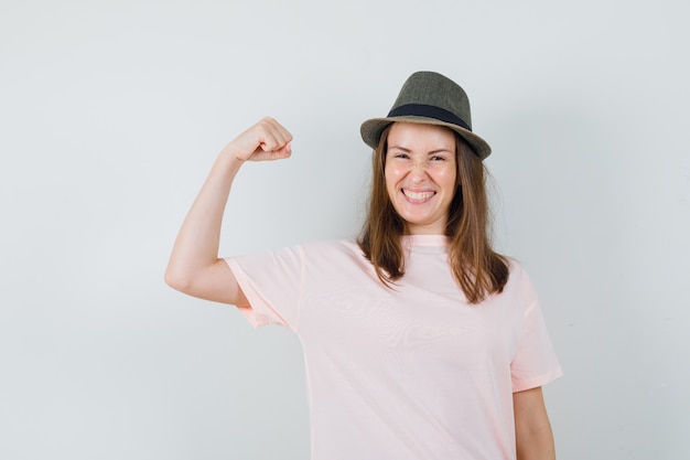 Chica joven mostrando gesto de ganador con sombrero de camiseta rosa y mirando dichoso