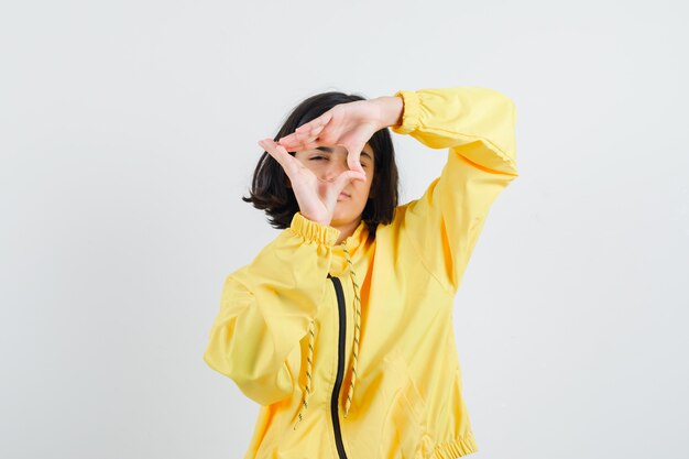 Chica joven mostrando gesto de marco en chaqueta de bombardero amarilla y mirando serio.