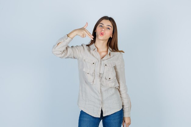 Chica joven mostrando gesto de pistola en camisa beige, jeans y luciendo lindo. vista frontal.