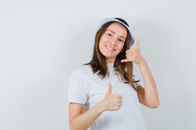 Chica joven mostrando gesto de teléfono con el pulgar hacia arriba en camiseta blanca, sombrero y mirando alegre, vista frontal.