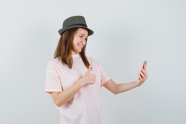 Chica joven mostrando el pulgar hacia arriba en el chat de video en camiseta rosa, sombrero y mirando alegre, vista frontal.