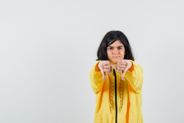 Chica joven mostrando los pulgares hacia abajo con ambas manos en chaqueta de bombardero amarilla y mirando disgustado.
