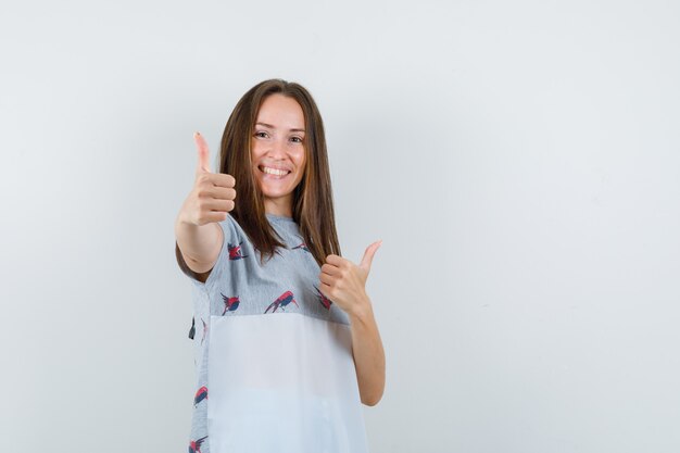 Chica joven mostrando los pulgares para arriba en camiseta y mirando feliz, vista frontal.