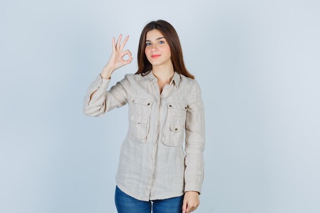 Chica joven mostrando signo ok en camisa beige, jeans y mirando alegre. vista frontal.