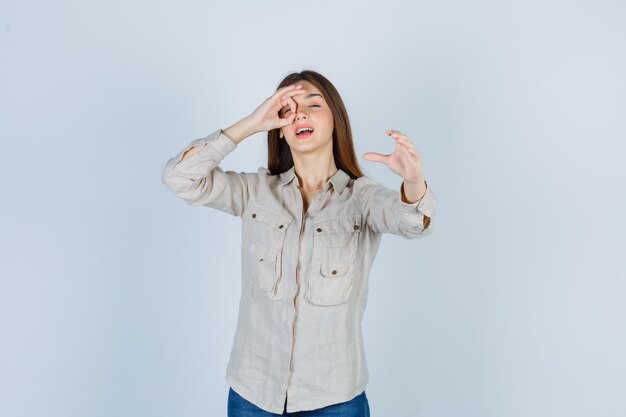 Chica joven mostrando el signo de ok en el ojo, estirando la mano hacia en camisa beige, jeans y luciendo linda. vista frontal.