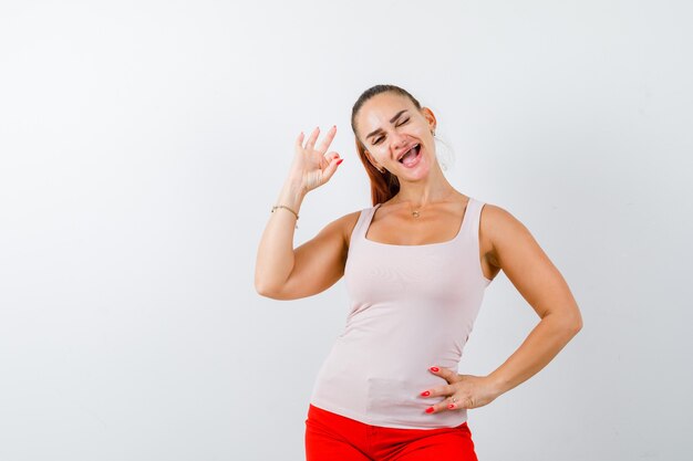 Chica joven mostrando el signo de ok, sosteniendo la mano en la cintura en top beige y pantalones rojos y mirando alegre, vista frontal.