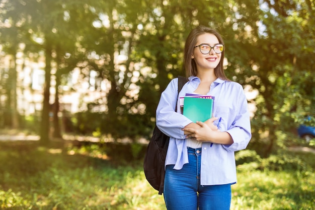 Chica joven muy atractiva con libros de pie y sonriendo en el parque