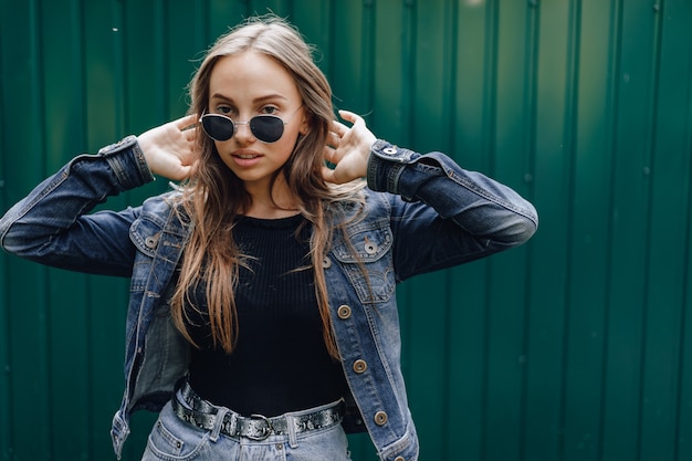 Chica joven muy atractiva en ropa de mezclilla en gafas sobre un fondo verde oscuro simple con lugar vacío para el texto