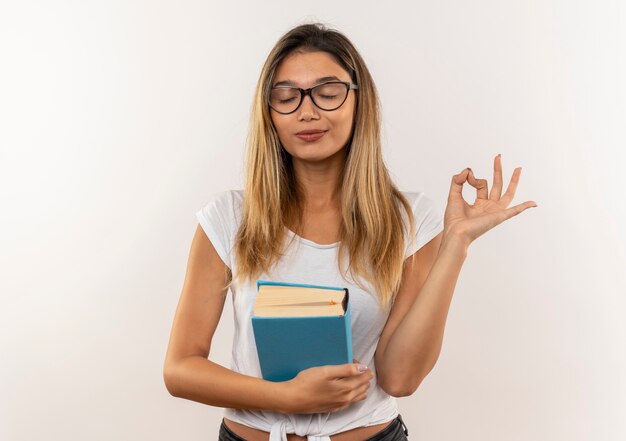 Chica joven y pacífica estudiante bonita con gafas y bolsa trasera sosteniendo el libro y haciendo el signo de ok con los ojos cerrados aislado sobre fondo blanco con espacio de copia