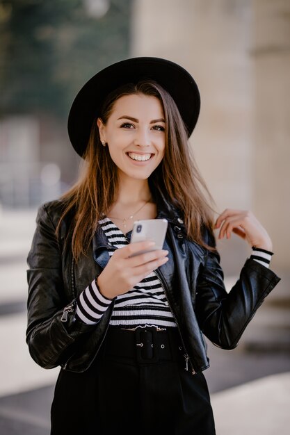 Chica joven de pelo castaño con una chaqueta de cuero, sombrero negro en el paseo de la ciudad posando con teléfono móvil