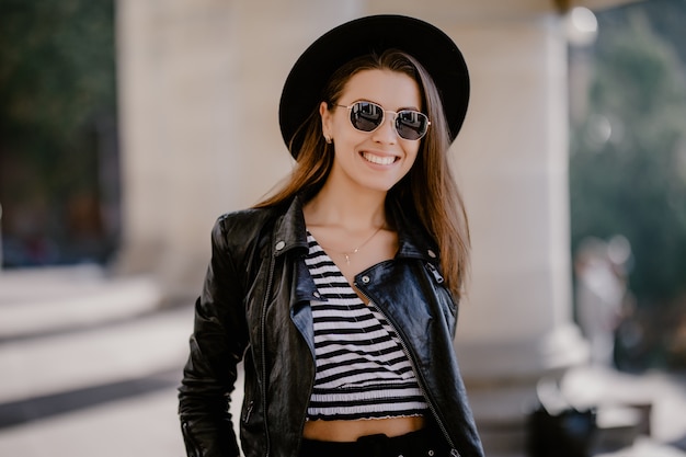 Chica joven de pelo castaño con una chaqueta de cuero, sombrero negro en el paseo de la ciudad