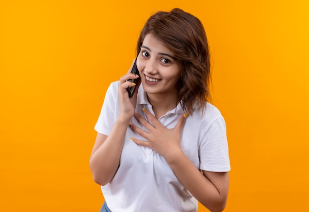 Chica joven con pelo corto con camisa polo blanca sintiéndose agradecida y feliz mientras habla por teléfono móvil