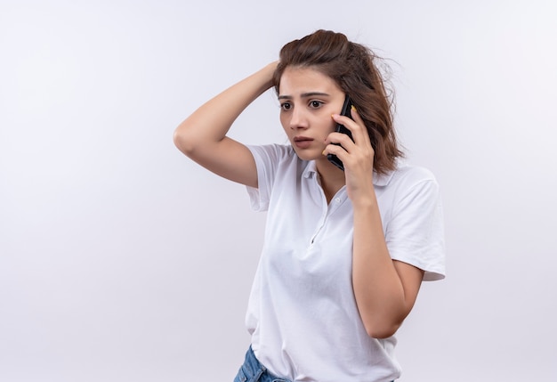 Chica joven con pelo corto vistiendo camisa polo blanca mirando confundido y muy ansioso mientras habla por teléfono móvil