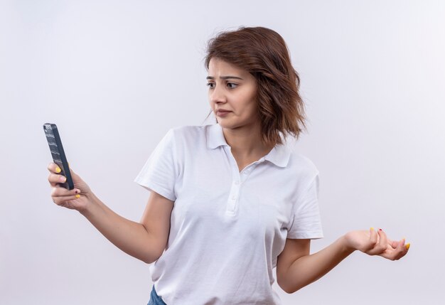 Chica joven con pelo corto vistiendo camisa polo blanca mirando la pantalla de su móvil con expresión confusa