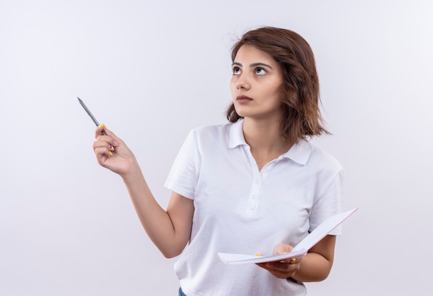 Chica joven con pelo corto vistiendo camisa polo blanca sosteniendo cuaderno y bolígrafo mirando a un lado con expresión pensativa