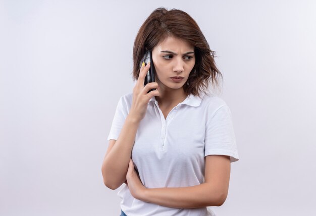 Chica joven con pelo corto vistiendo polo blanco hablando por teléfono móvil con el ceño fruncido