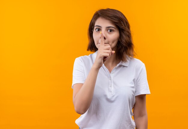 Chica joven con pelo corto vistiendo polo blanco haciendo gesto de silencio con el dedo en los labios