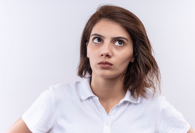 Chica joven con pelo corto vistiendo polo blanco mirando hacia arriba con expresión sospechosa