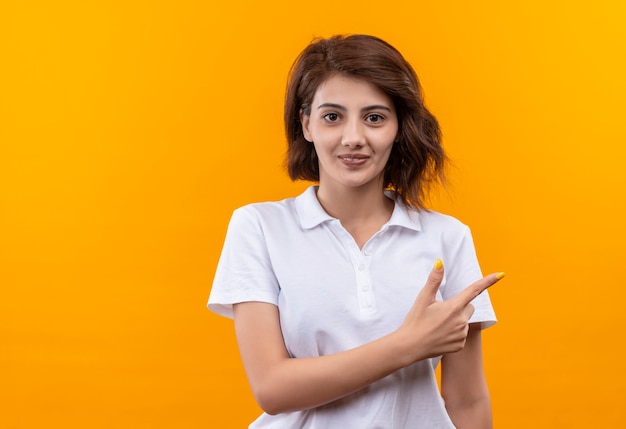 Chica joven con pelo corto vistiendo polo blanco mirando confiado apuntando con el dedo índice hacia el lado