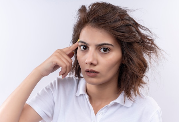 Chica joven con pelo corto vistiendo polo blanco mirando confundido y preocupado apuntando su sien