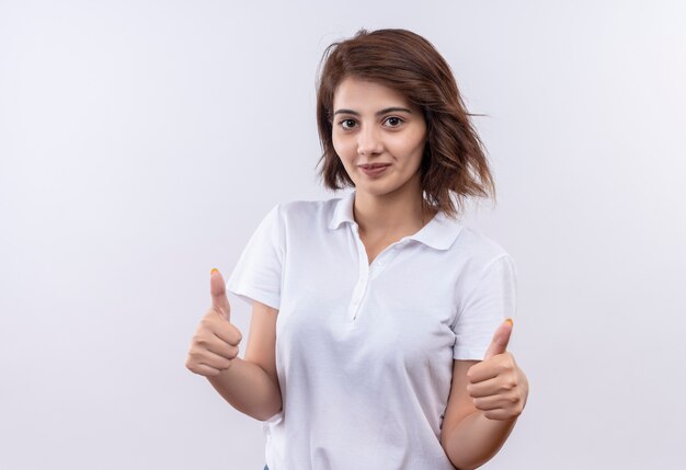 Chica joven con pelo corto vistiendo polo blanco sonriendo amable mostrando los pulgares hacia arriba con ambas manos