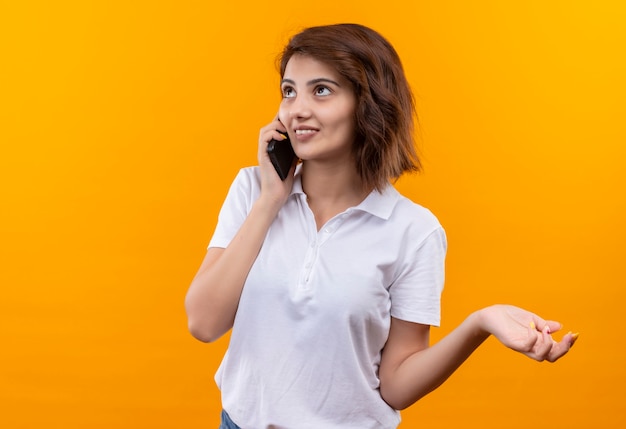 Chica joven con pelo corto vistiendo polo blanco sonriendo mientras habla por teléfono móvil