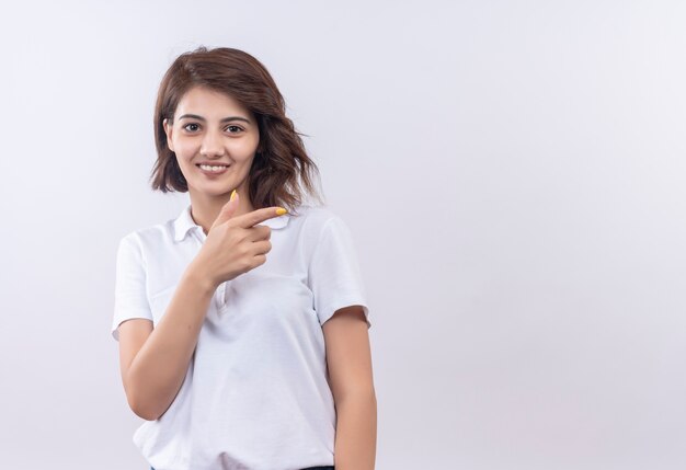 Chica joven con el pelo corto vistiendo polo blanco sonriendo positivo y feliz apuntando con el dedo hacia el lado