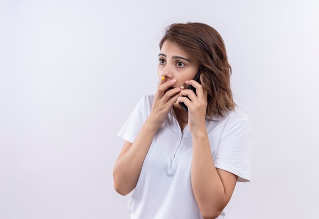 Chica joven con pelo corto vistiendo polo blanco sorprendido mientras habla por teléfono móvil cubriendo la boca con la mano