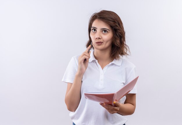 Chica joven con pelo corto vistiendo polo blanco sosteniendo el cuaderno y la pluma mirando a un lado con expresión pensativa, pensando