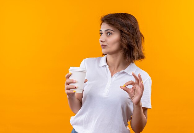 Chica joven con pelo corto vistiendo polo blanco sosteniendo la taza de café sonriendo alegremente mostrando signo ok