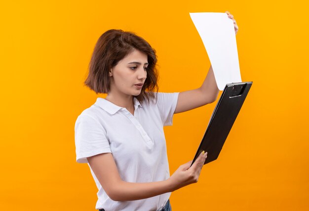 Chica joven con pelo corto vistiendo polo blanco sujetando el tablero de clip mirando páginas en blanco con cara seria