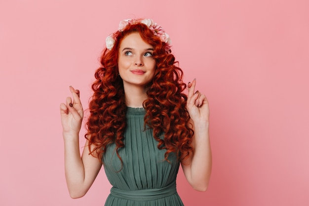 Foto gratuita chica joven con pelo rojo y flores en la cabeza cruza los dedos. retrato de mujer de ojos azules en vestido verde en espacio rosa.