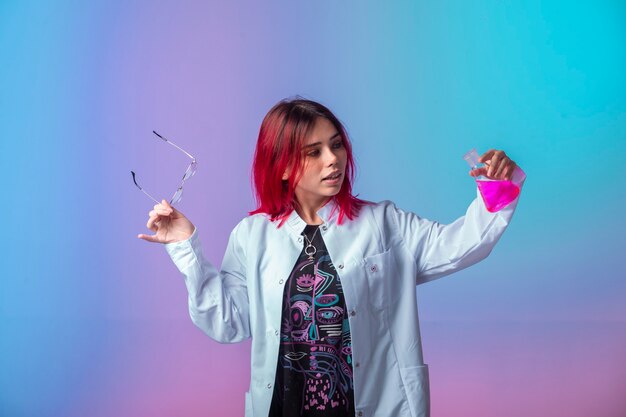 Chica joven con pelos rosados sosteniendo un matraz químico y mirando con atención.